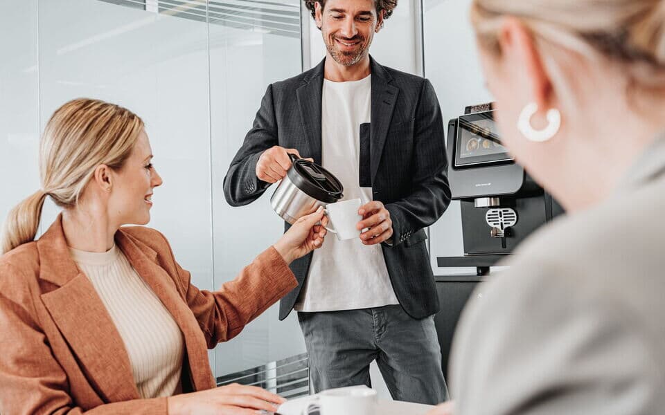 Ein Mann gießt Kaffee aus einer Kanne in eine Tasse einer Kollegin, während sie an einem Tisch in einem modernen Büro sitzt. Eine weitere Kollegin sitzt im Vordergrund, während im Hintergrund ein Kaffeevollautomat zu sehen ist.