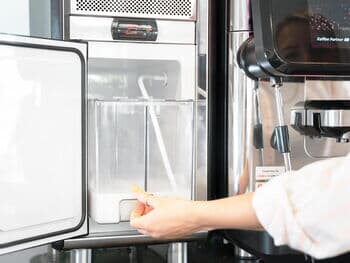 Nahaufnahme eines Kaffeevollautomaten von Kaffee Partner mit angeschlossenem Milchschlauch und Milchkühler, Hand zeigt auf den Milchkühler