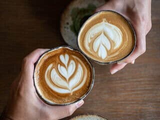 zwei kaffeebecher aus keramik in zwei händen gegenüber mit latte art herz oben drauf