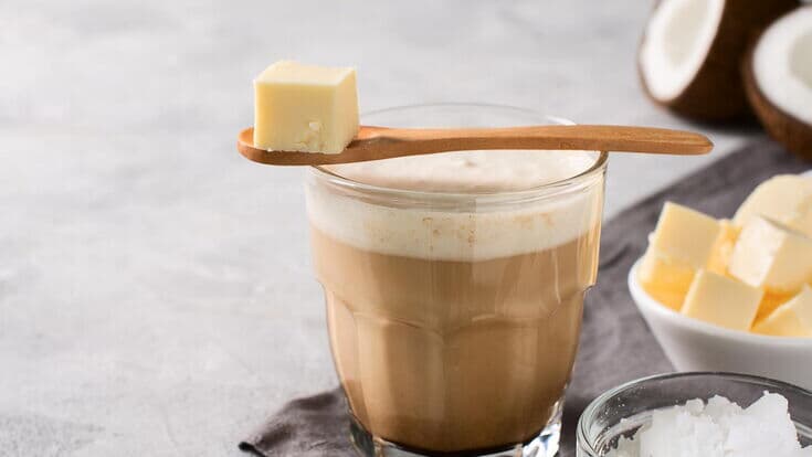 Ein Glas Bulletproof Coffee mit cremiger Schaumkrone, davor Butterwürfel, MCT-Öl und eine Kokosnusshälften.