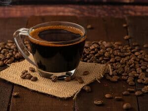 Glastasse mit schwarzem Kaffee umgeben von Kaffeebohnen auf Jutestoff platziert auf rustikalem Holztisch