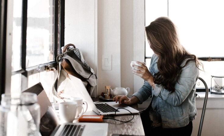 Eine Frau mit langen Haaren sitzt im Büro an einem Laptop und hält eine Tasse Kaffee in der Hand