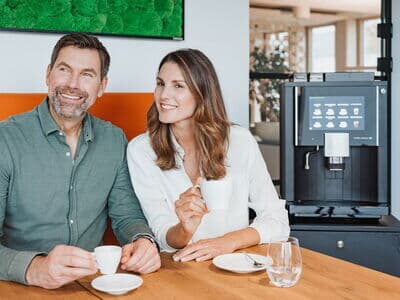 Lächelnde Frau und Mann genießen gemeinsam Kaffee im Büro, zubereitet mit einem Kaffeevollautomaten der BARISTA-Serie von Kaffee Partner.
