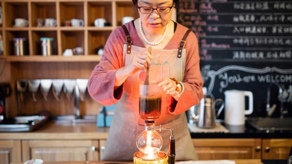 Eine asiatische Frau rührt mit einem Holzlöffel den Kaffee in einem Syphon um