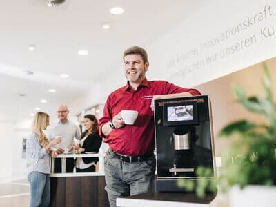 Servicemitarbeiter steht neben einem Kaffeeautomaten und trinkt Kaffee.