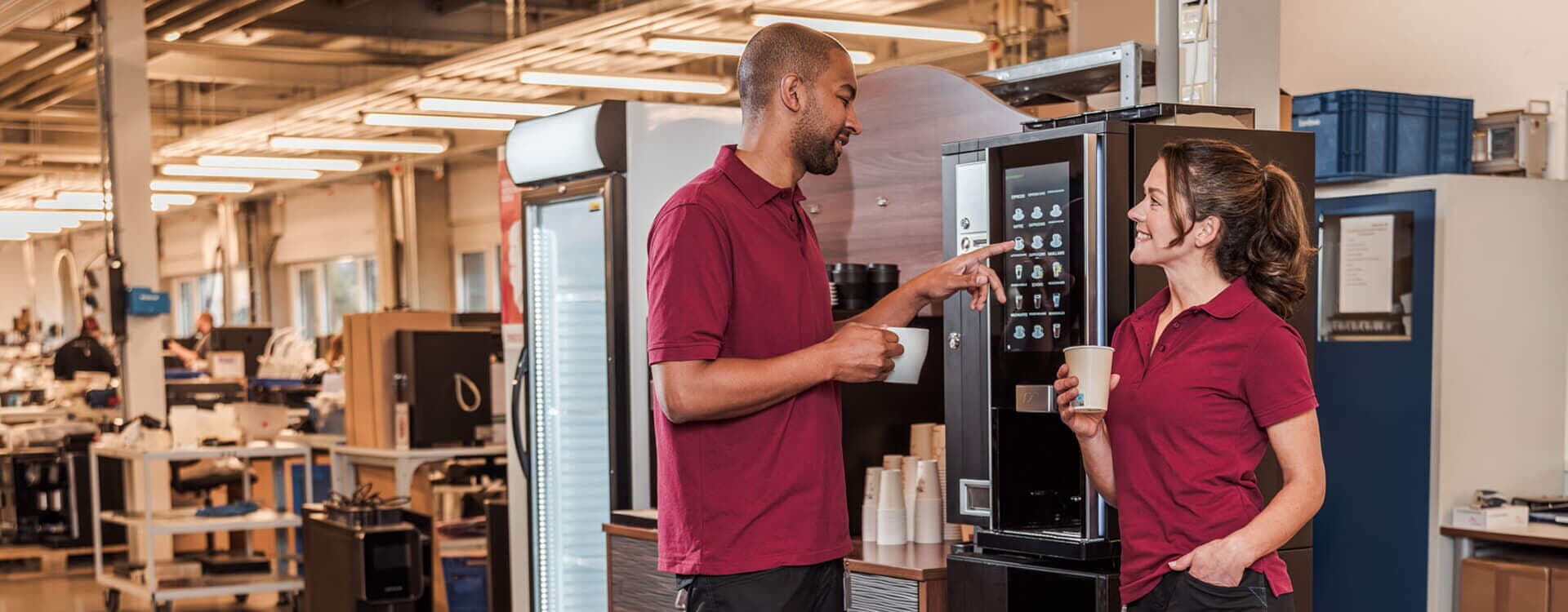 Zwei Mitarbeiter trinken Kaffee neben Kaffeevollautomat in einer Werkstatt.
