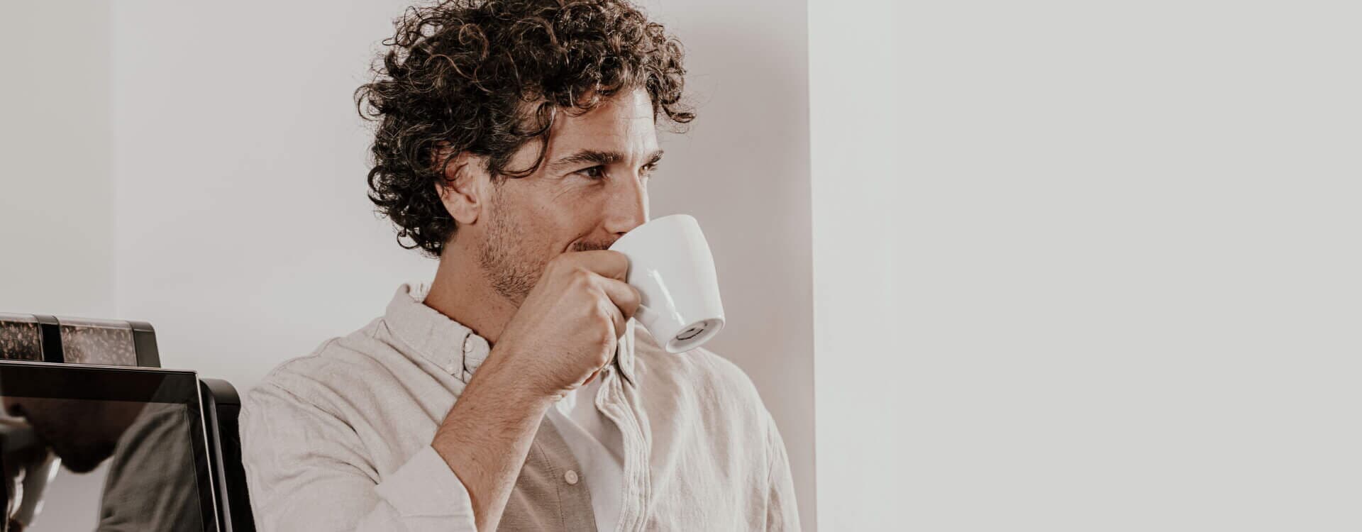 Ein Mann mit lockigem Haar genießt eine Tasse Kaffee, während er neben einem Kaffeevollautomaten von Kaffee Partner steht.