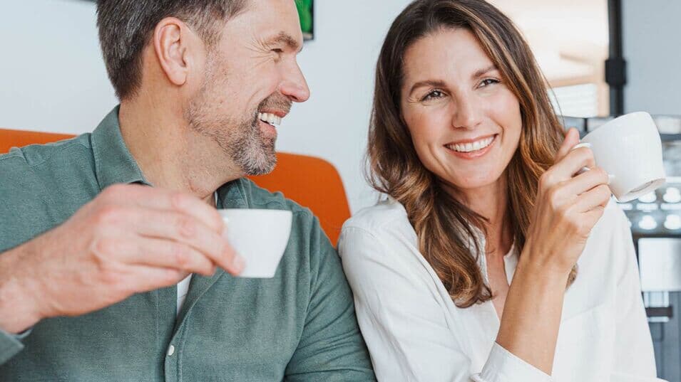 Mann und Frau sitzen auf orangenem Sofa und einem Kaffeevollautomaten im Hintergrund, trinken Kaffee und lachen